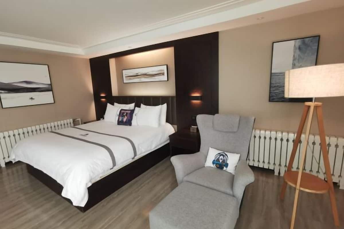 A hotel room with a large bed, a grey armchair, a standing lamp, and artwork on the walls.