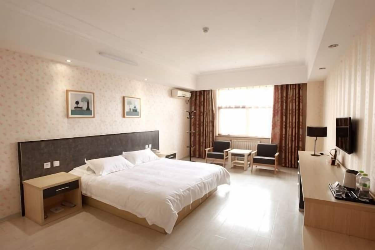 A hotel room with a large bed, bedside tables, a desk, a chair, a TV, and a window with curtains.