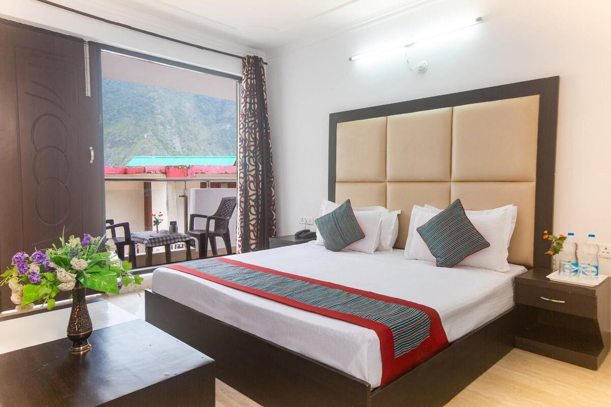 A hotel room with a large bed, a bedside table with a vase of flowers, a window with a mountain view, and a desk with a chair.