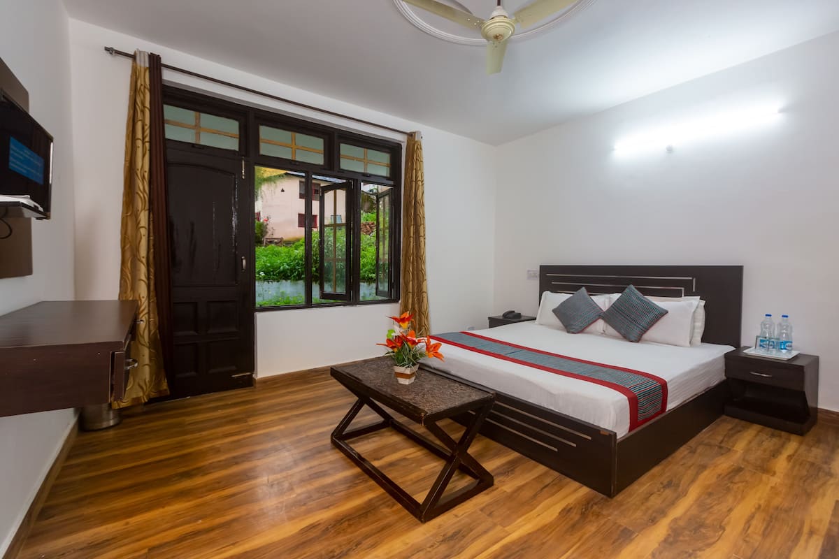 A hotel room with a wooden floor, a bed with a red and blue striped bedspread, a nightstand with water bottles, and a window with curtains.