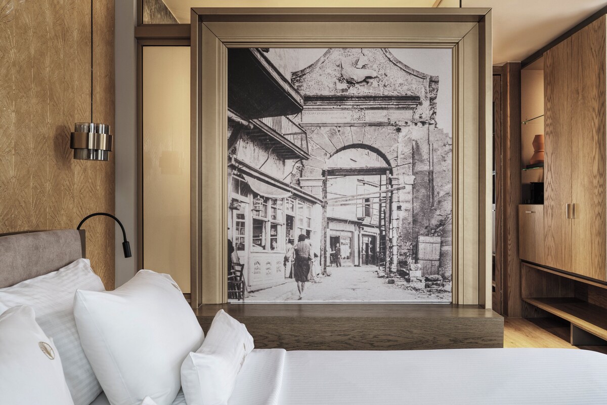 Una habitación de hotel moderna con una fotografía grande en blanco y negro enmarcada de una escena callejera, una cama bien hecha y un armario de madera.