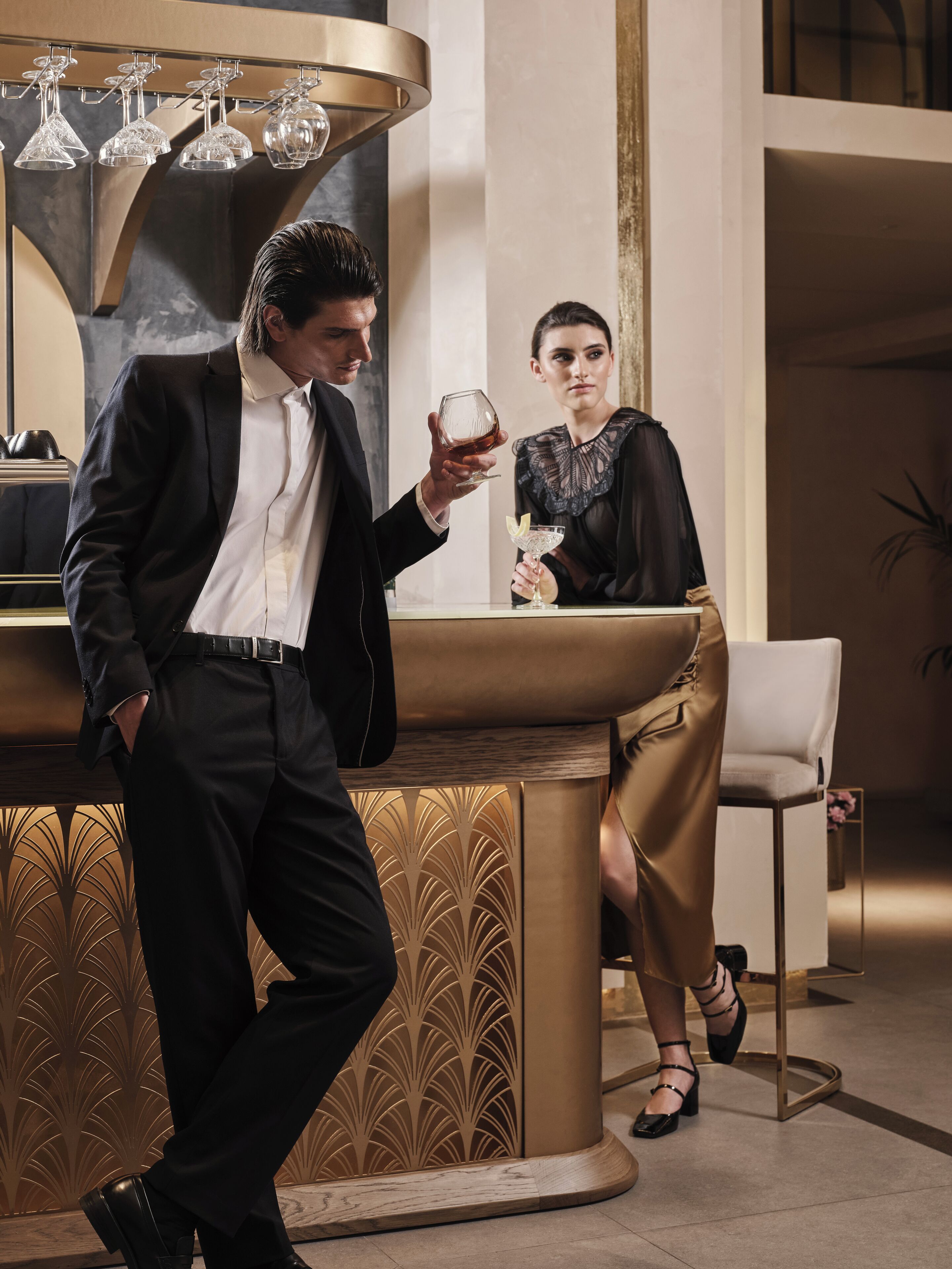 A man in a black suit holding a drink at a bar with a woman in a black dress seated behind it.
