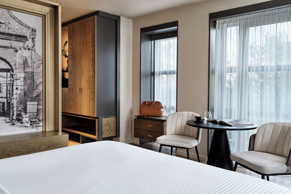 Una habitación de hotel con una cama, dos sillas, una mesita, un armario y un ventanal con cortinas.