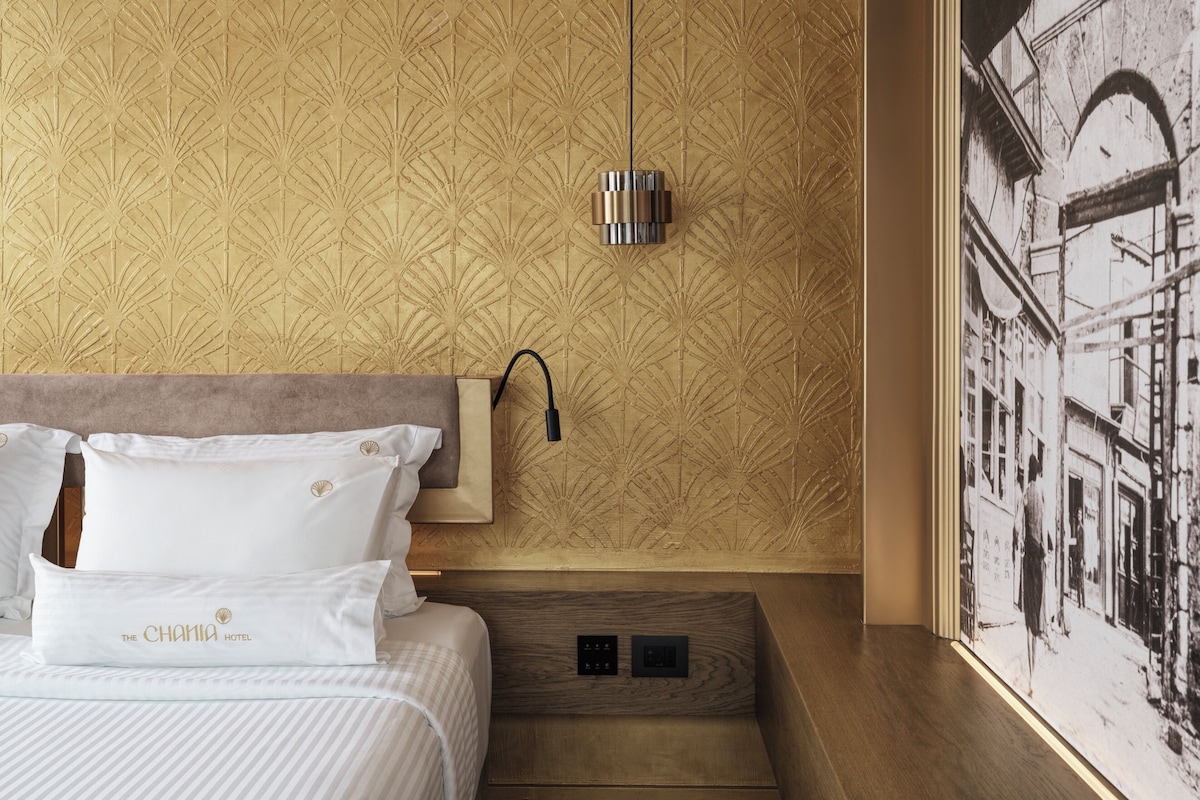 Una habitación de hotel con papel tapiz estampado, una cama con ropa de cama blanca, una mesita de noche y una lámpara fijada a la pared.