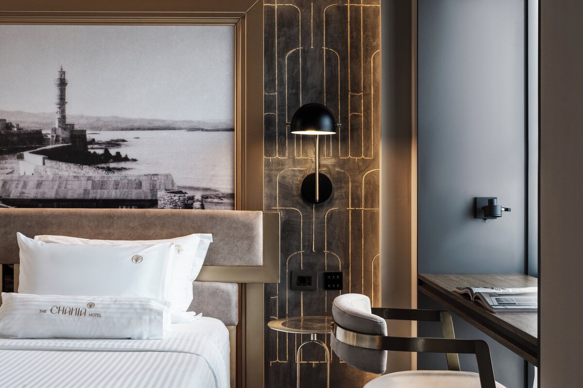 Una habitación de hotel con una cama, una mesita de noche, una silla, un escritorio con una lámpara y una lámpara fijada a la pared.