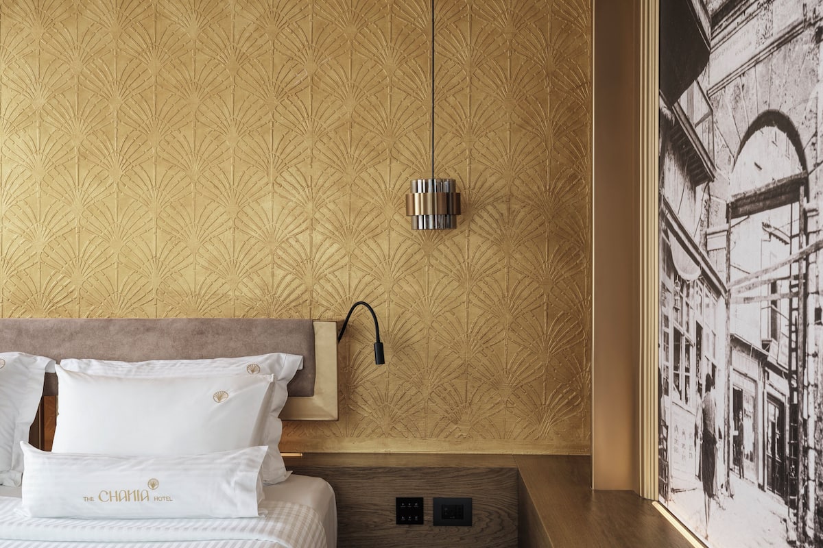 Une chambre d’hôtel avec un papier peint à motifs, un lit avec une literie blanche, une table de chevet et une lampe fixée au mur.