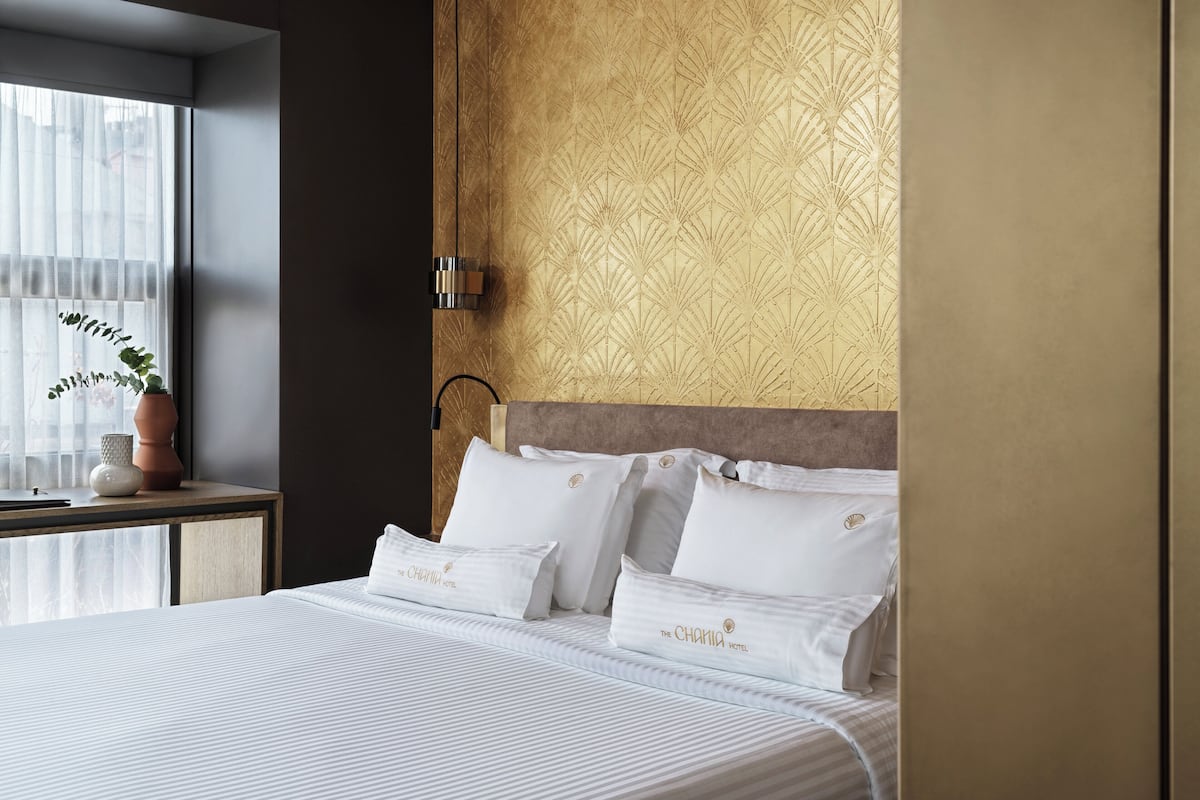 Une chambre d’hôtel avec un lit, une table de chevet ornée d’une plante, un mur décoré d’un motif et une fenêtre avec des rideaux légers.