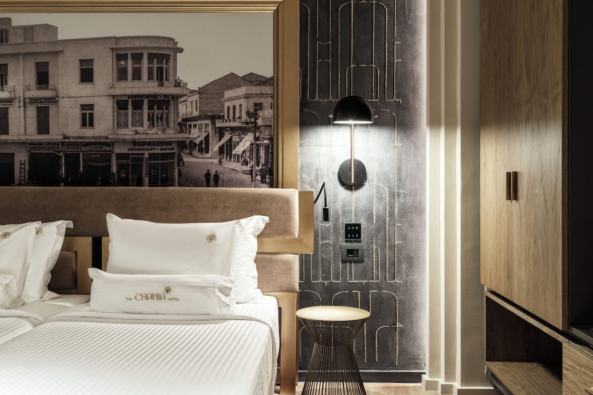 Une chambre d’hôtel avec un grand lit, une lampe fixée au mur et une fresque murale représentant un paysage urbain d’époque.