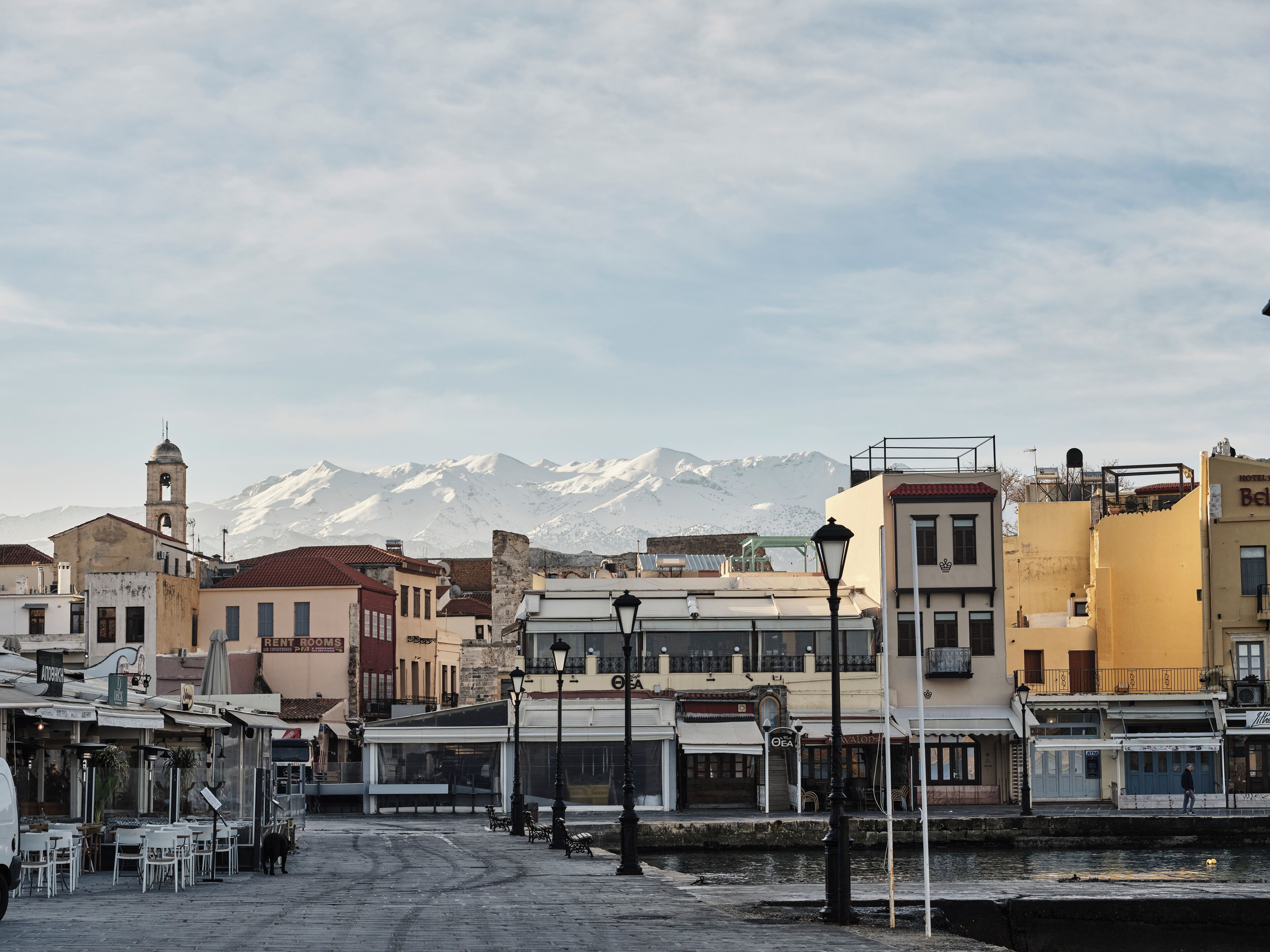 A waterfront street with buildings, a bell tower, and snow-capped mountains in the background.