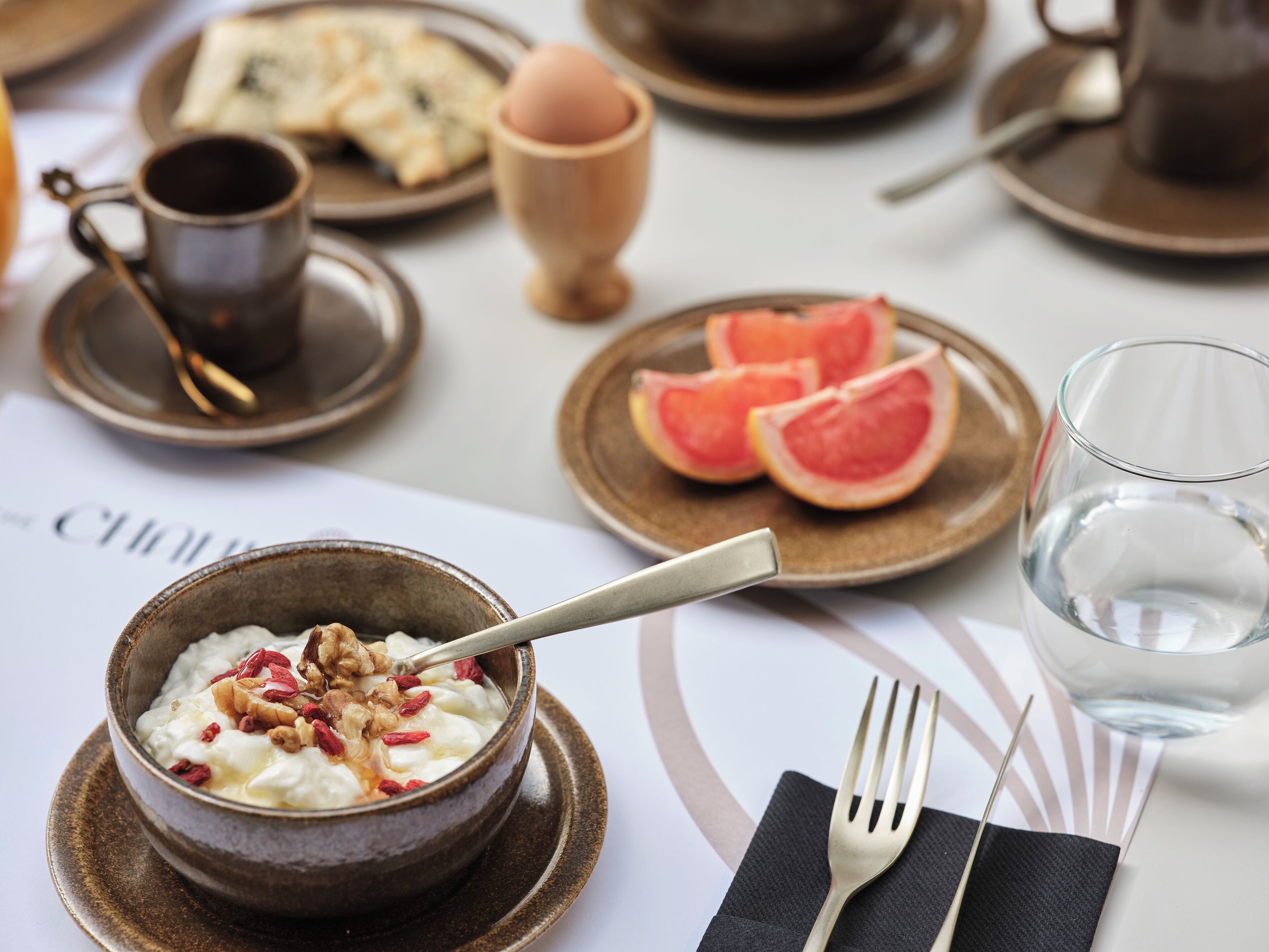 Une table mise pour le petit-déjeuner avec un bol de yaourt garni de noix et de fruits secs, une assiette de pamplemousse et un verre d’eau.