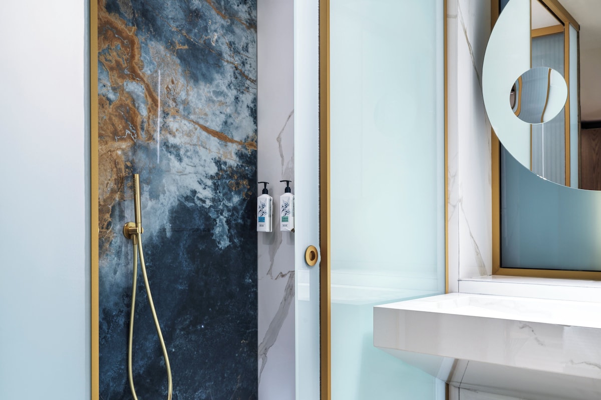 Un baño con pared de mármol, cabezal de ducha dorado y espejo con marco circular.