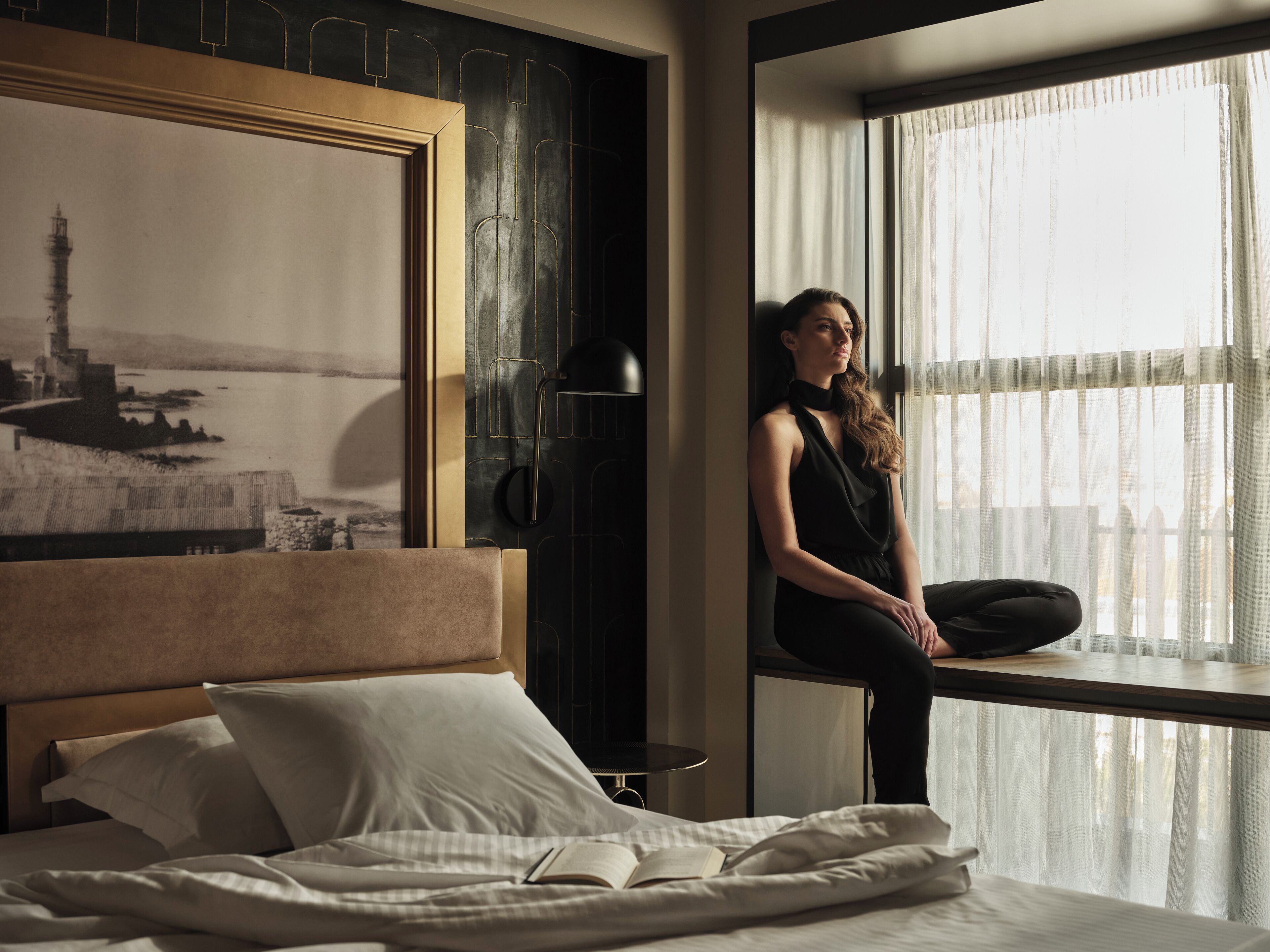 A hotel room with a large bed, a window with sheer curtains, a framed picture on the wall, and a black lamp.