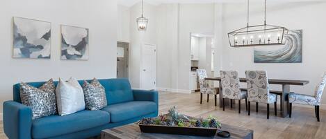 Una sala de estar moderna con un sofá azul, una mesa de centro con objetos decorativos, un comedor con sillas estampadas y arte abstracto en la pared.