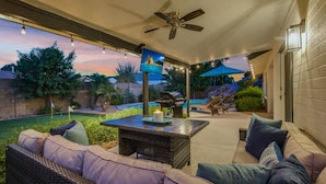 Un patio al aire libre con ventilador de techo, una mesa con una vela y vistas a un jardín trasero con piscina y zona de barbacoa.