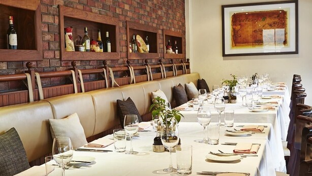 Una larga mesa de comedor dispuesta con copas de vino, platos y cubiertos, flanqueada por cabinas de cuero y una pared de ladrillo con botellas de vino.