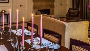 Mesa de comedor dispuesta para seis personas, con velas rosadas, chimenea y alfombra estampada.