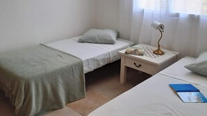 Ein kleines, sauberes Zimmer mit zwei Einzelbetten, einem Nachttisch mit Lampe und einem Fenster mit durchsichtigen Vorhängen.
