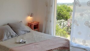 Ein Schlafzimmer mit Bett, Kissen, einem Nachttisch mit Lampe und Blick auf Grün durch ein Fenster.