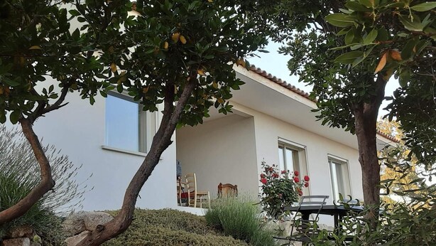 Ein modernes Haus mit weißer Fassade, einem Balkon mit Tisch und Stühlen sowie einem Garten mit verschiedenen Pflanzen und einem Baum.