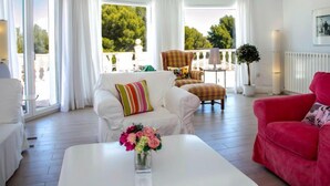A bright living room with a white sofa, a red armchair, and a coffee table with a vase of flowers. There are large windows with curtains, a balcony with a chair, and a radiator. - Villa Polonia Mouche by Interhome (Dénia)
