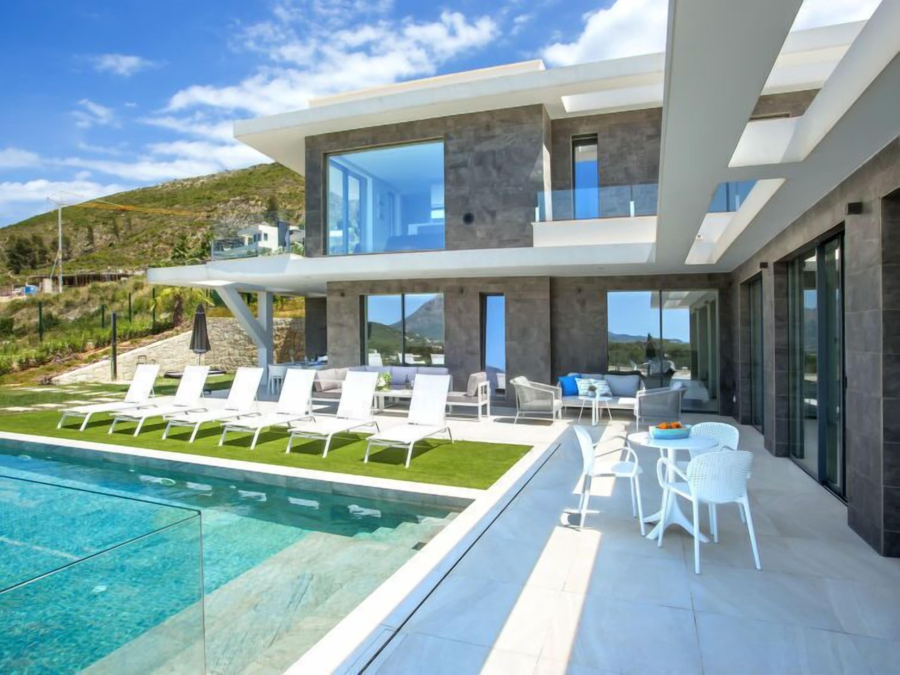 A modern two-story house with a swimming pool, outdoor seating, and a view of the surrounding landscape.