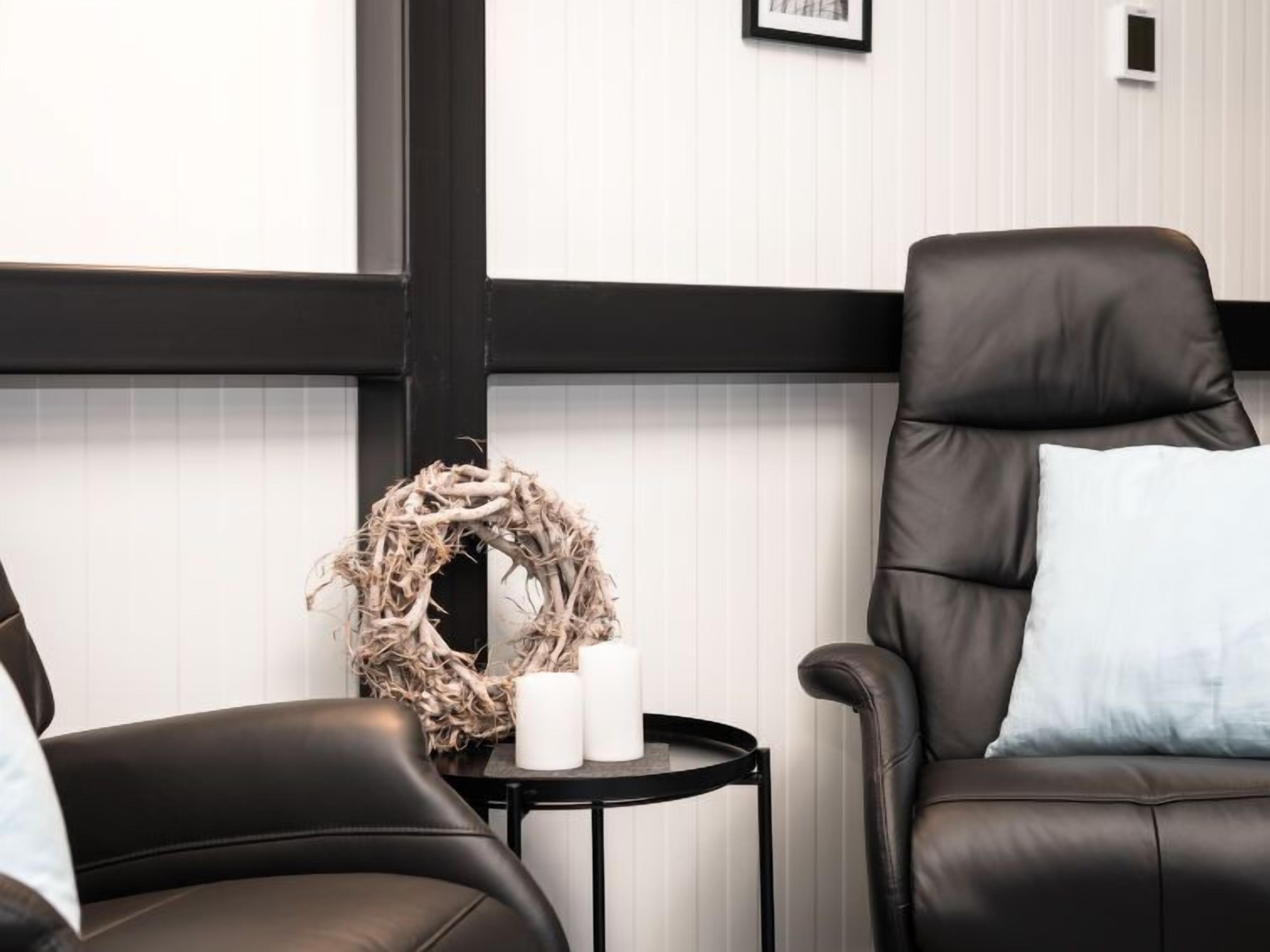 Une pièce avec un fauteuil en cuir noir, une petite table ronde ornée d’une guirlande et de bougies, et un mur blanc avec des lignes verticales.