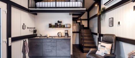 Compact living space with a central kitchen, seating area, and staircase leading to a balcony.