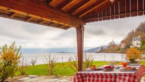 Tavolo apparecchiato all'aperto con tovaglia a quadretti, con vista su un lago e una casa sullo sfondo.