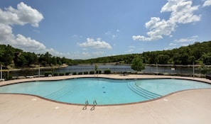 A large, empty swimming pool with a clear blue sky and a forested hill in the background.