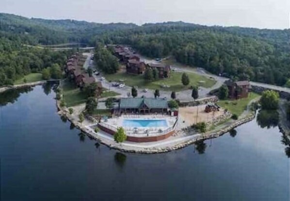 Vue aérienne d’un complexe hôtelier au bord d’un lac, avec une piscine, plusieurs bâtiments et une végétation luxuriante.