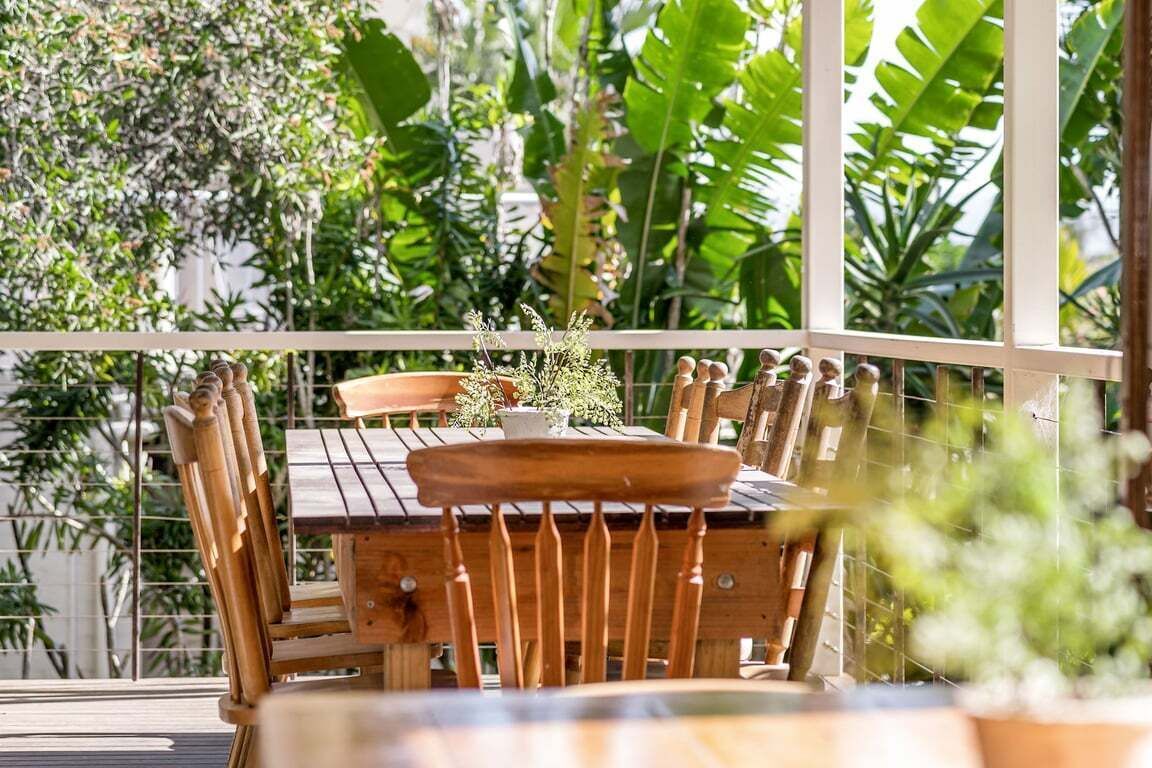 An outdoor wooden table with chairs, surrounded by a white fence, with lush green foliage in the background.