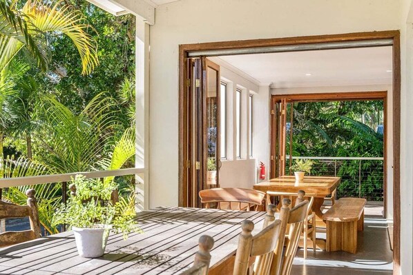 A wooden dining table with chairs, a potted plant, and a view of lush greenery through an open door.