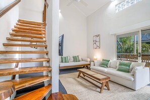 Una escalera con escalones de madera, un sofá blanco con cojines verdes, una mesa de centro de madera y un ventanal con vistas a zonas verdes.