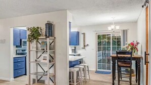 Cocina moderna con armarios azules, encimera blanca y estufa integrada. Cuenta con un comedor con mesa y sillas de madera, y un amplio ventanal con vistas al exterior.
