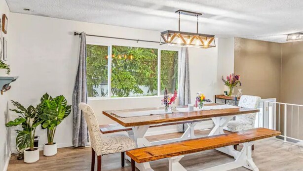 Un comedor con mesa y bancos de madera, ventanas amplias con cortinas y plantas en macetas.