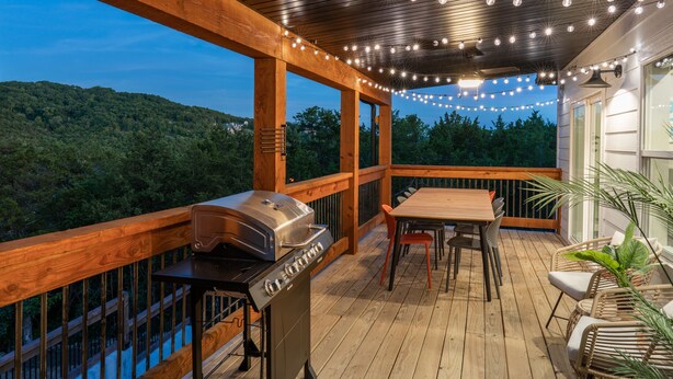 Una terraza de madera con una parrilla, mesa, sillas y luces colgantes.