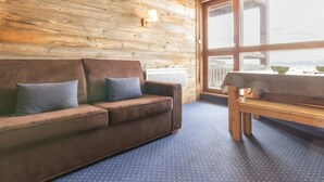 A cozy room with a wooden wall, a brown sofa with cushions, a wooden table, and a large window offering a view.