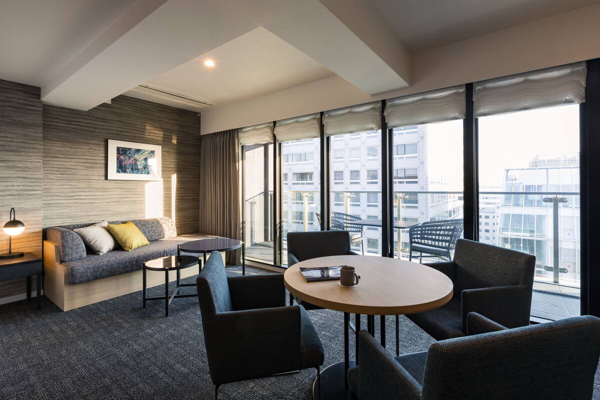 A modern hotel room with a sofa, chairs, a table, and large windows offering a city view.