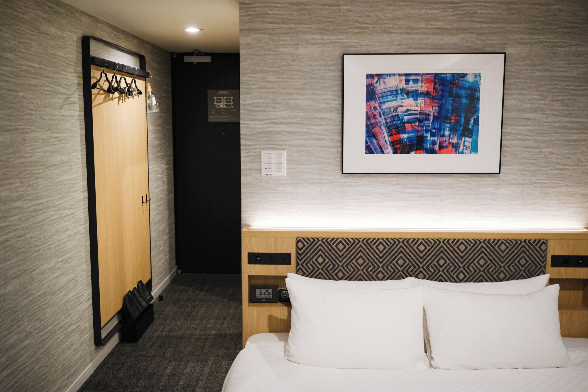 A hotel room with a bed, a headboard with a geometric pattern, a wall-mounted painting, a wardrobe, and a coat rack.