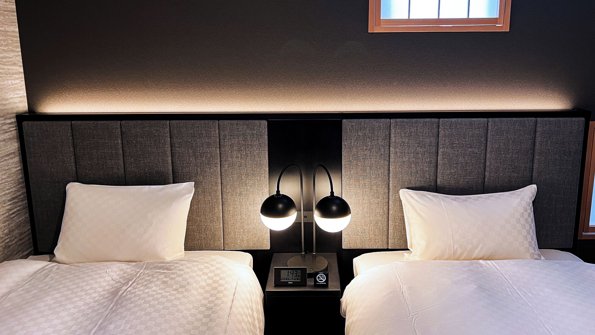 A hotel room with two beds, a nightstand with a clock and a no-smoking sign, and a window above the beds.