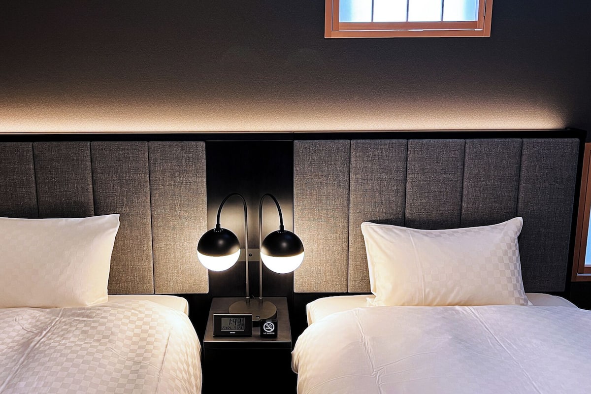 A hotel room with two beds, a nightstand with a clock and a no-smoking sign, and a window above the beds.