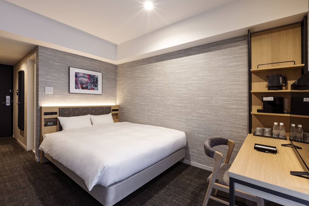 A modern hotel room with a large bed, a desk with a chair, and a built-in shelf with a coffee maker and bottles.