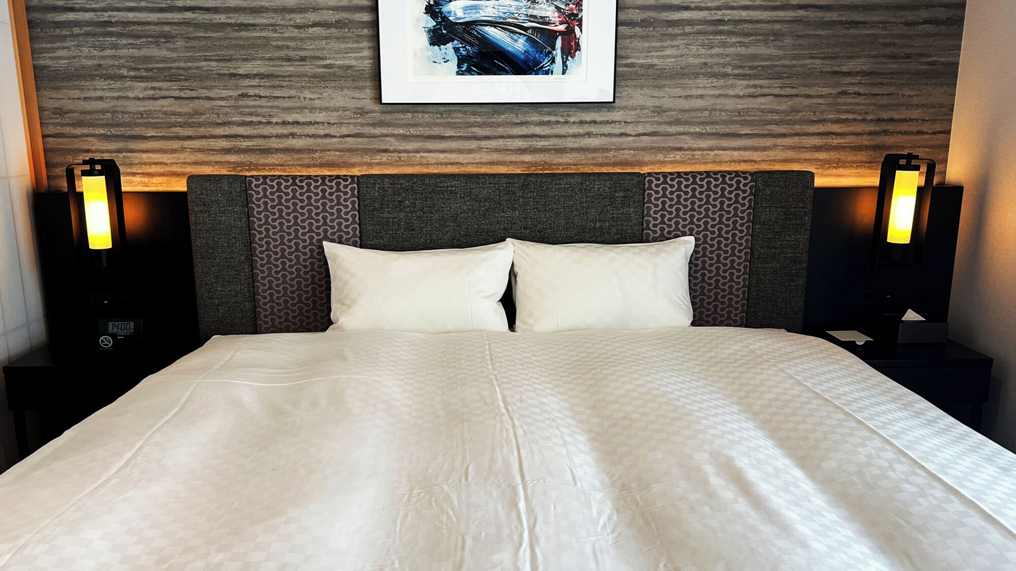 A neatly made bed with a patterned bedspread, a headboard with a geometric design, and two bedside lamps.