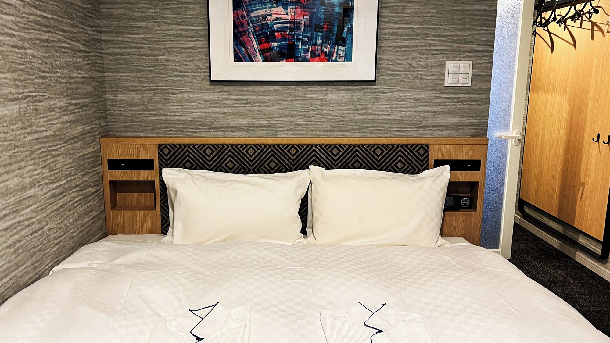 A hotel room with a checkered bedspread, a patterned headboard, a framed cityscape artwork, and a wooden wardrobe.
