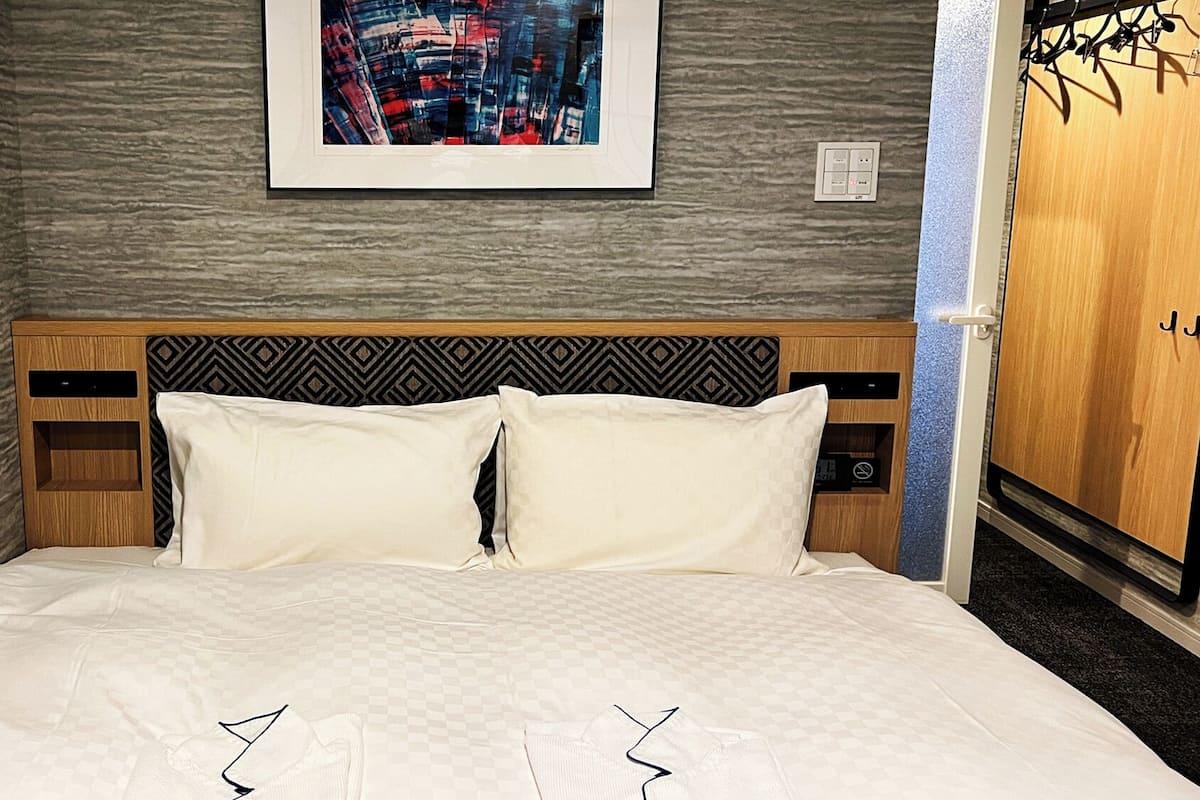 A hotel room with a checkered bedspread, a patterned headboard, a framed cityscape artwork, and a wooden wardrobe.