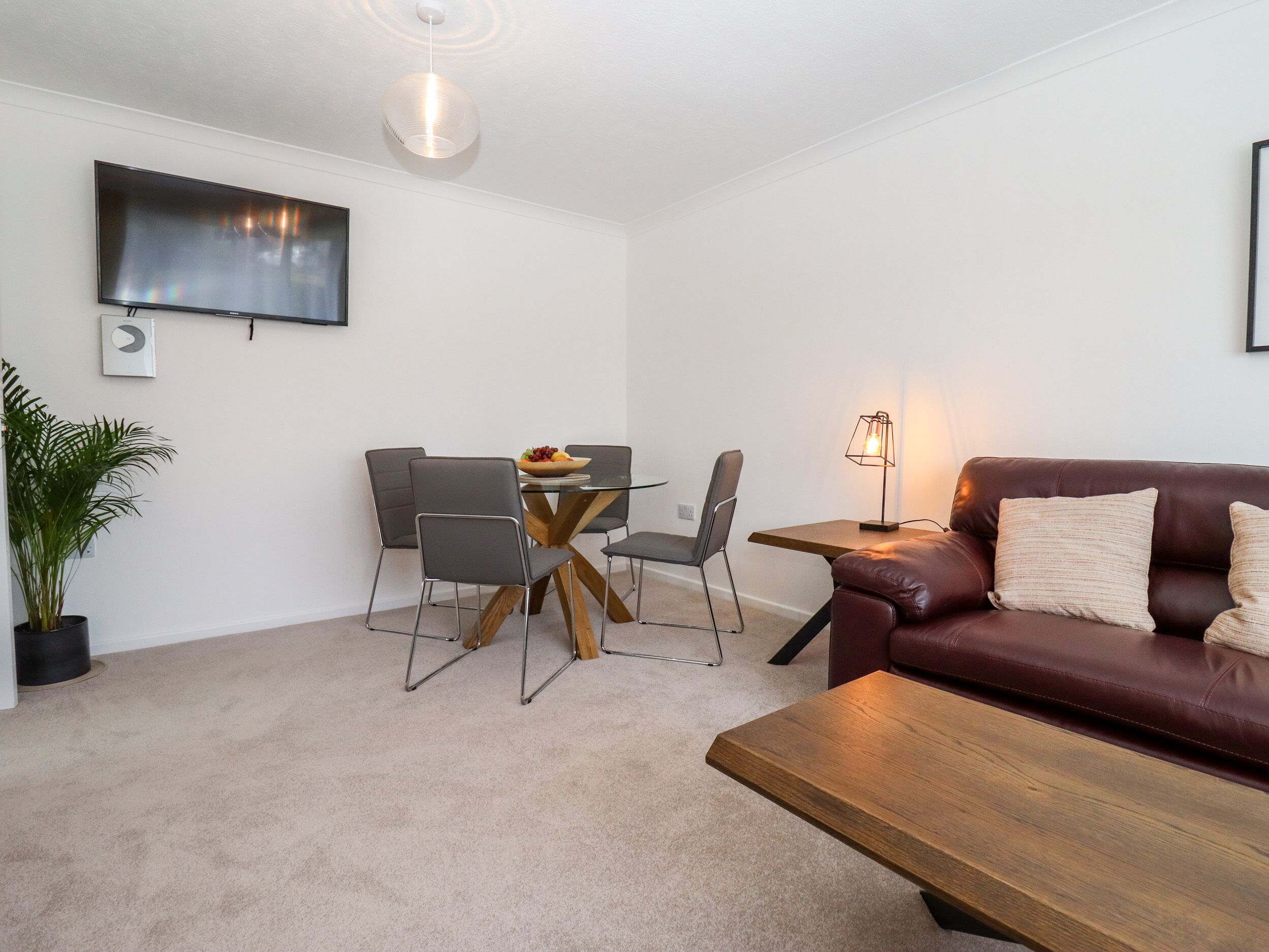 A modern living room with a brown leather sofa, a wooden coffee table, a dining table with four chairs, a TV mounted on the wall, and a potted plant.