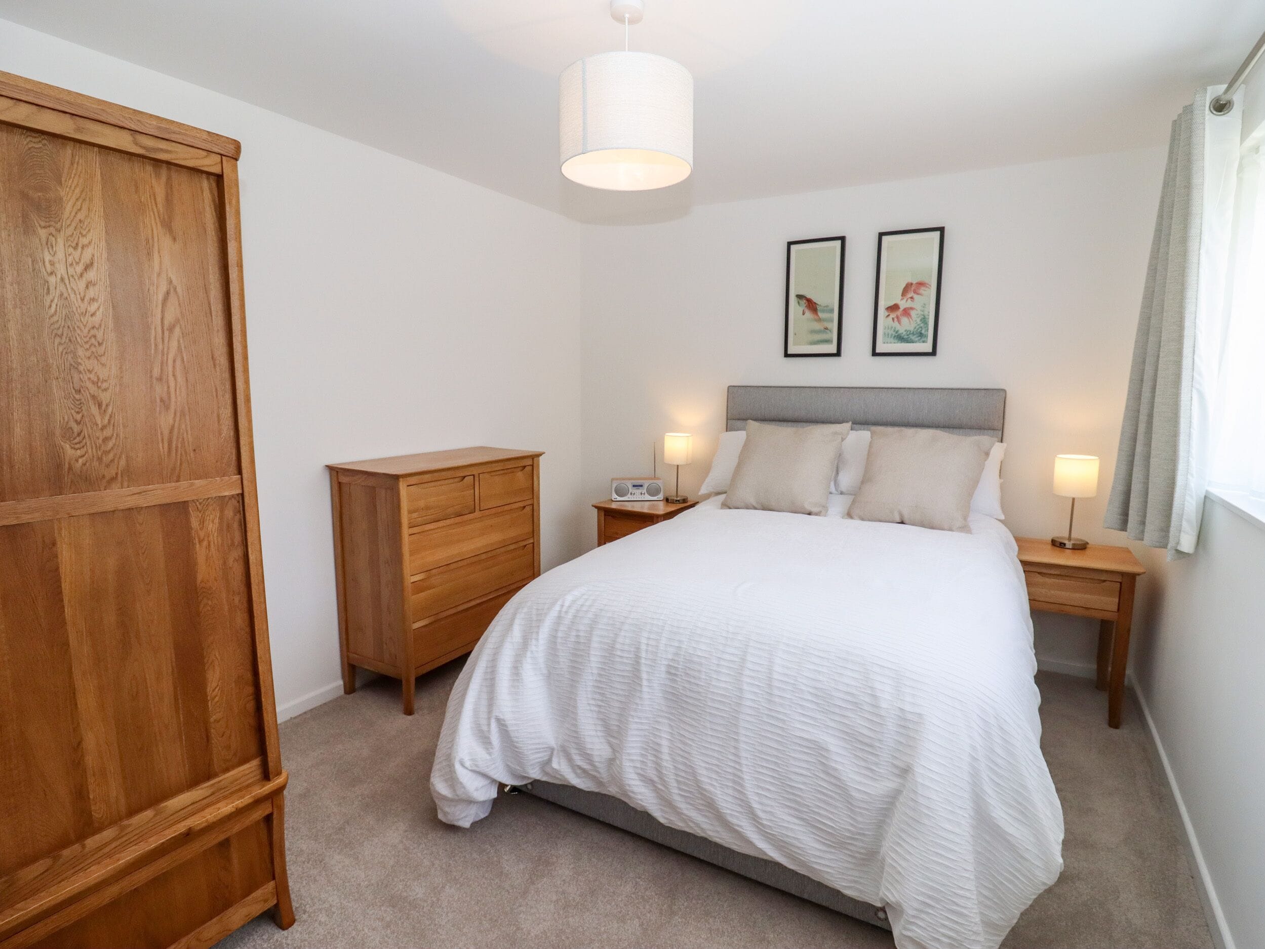 Un dormitorio con cama, armario de madera, mesitas de noche con lámparas y dos cuadros en la pared.