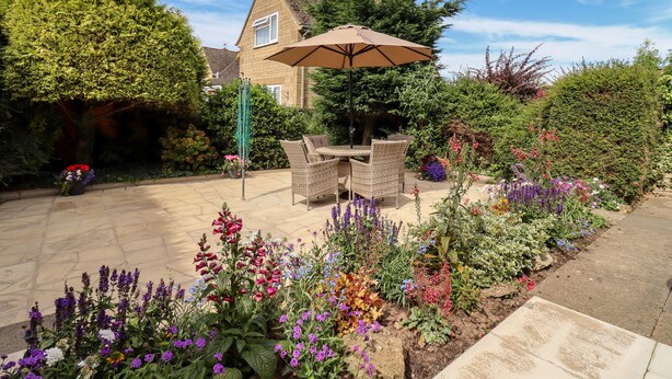 A patio area with a dining table and chairs, surrounded by a variety of colorful flowers and plants. - 3 Granbrook Lane (Chipping Campden)