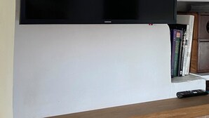 Seinälle asennettu litteänäyttöinen televisio, jonka yläpuolella on puinen hylly, jossa on kirjoja ja kaukosäädin.