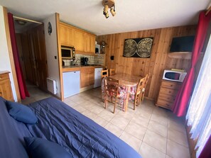 A compact room with a bed, a dining table with chairs, a kitchenette with a microwave, and a small cabinet.